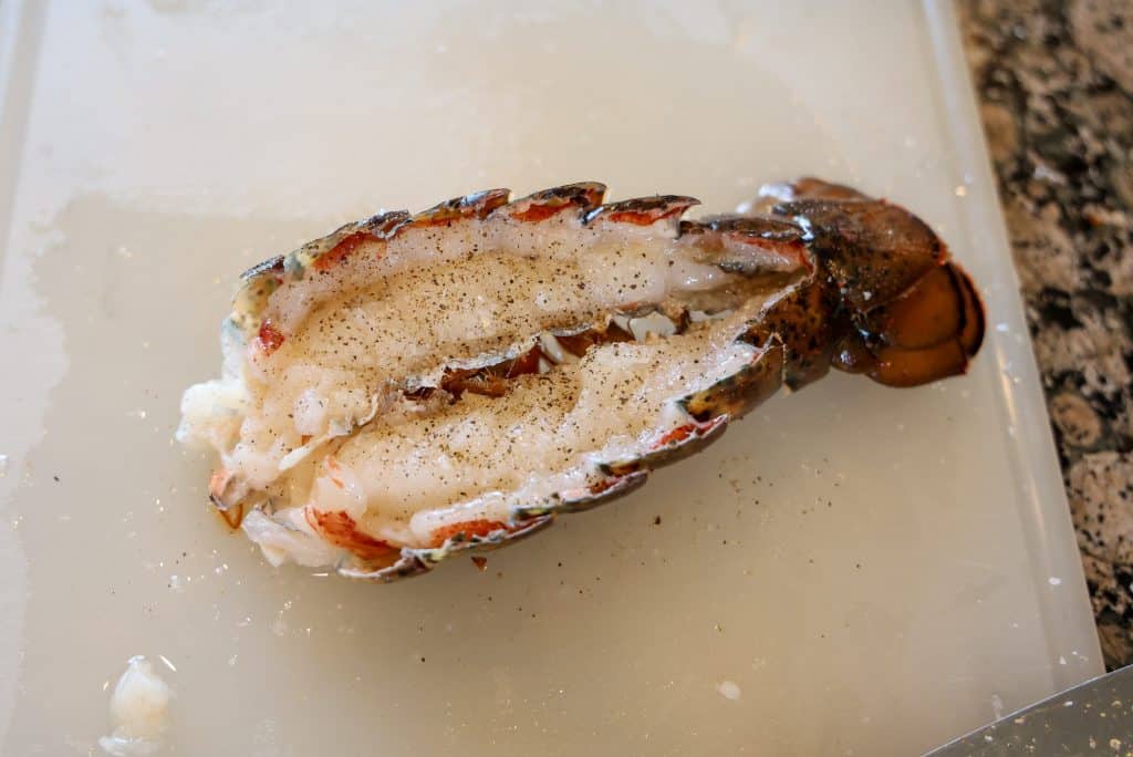 a halved uncooked lobster tail with salt and pepper sprinkled on top on a white cutting board