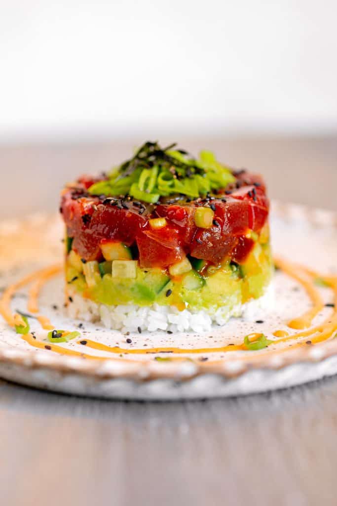 White plate with circle of spicy mayo around the rim. Stack of tuna, avocado, cucumber, and rice layered on top of plate.