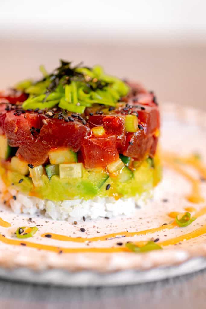 close up of a spicy tuna sushi stack with layers of diced cucumber, avocado, and rice on a white plate