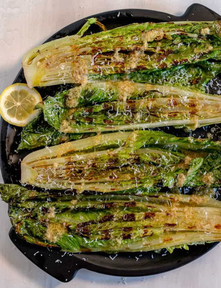Grilled heads of romaine on a black circle plate with sliced lemons surrounding plate