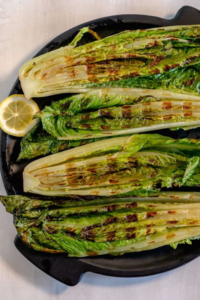 grilled romaine hearts on a black plate with a lemon slice on the side
