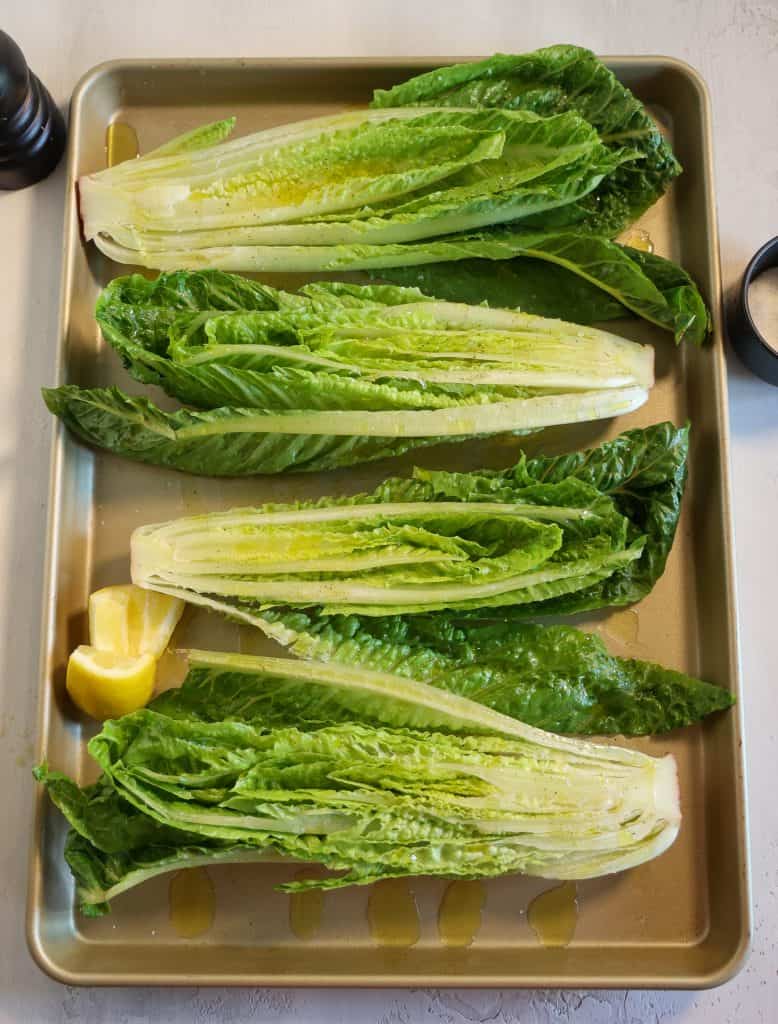Cut romaine hearts on a gold oven sheet with sliced lemon, pepper, and salt surrounding on the side
