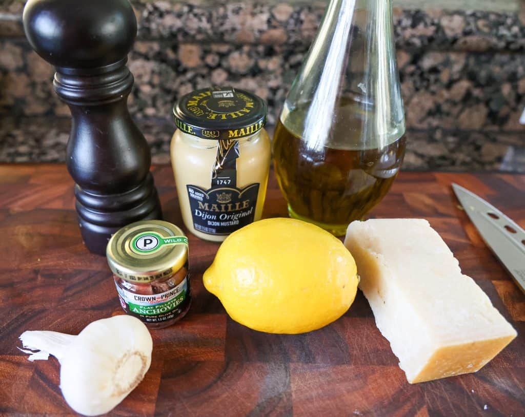 a lemon, block of cheese, anchovy filets, garlic, dijon mustard on a wooden cutting board