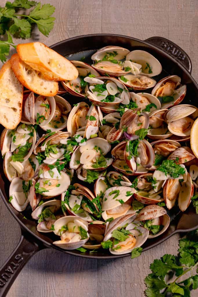 cast iron pan with open cooked clams topped with chopped cilantro, a lemon wedge on the right, on a grey wooden background surrounded by fresh sprigs of cilantro and three pieces of toasted bread in the corner