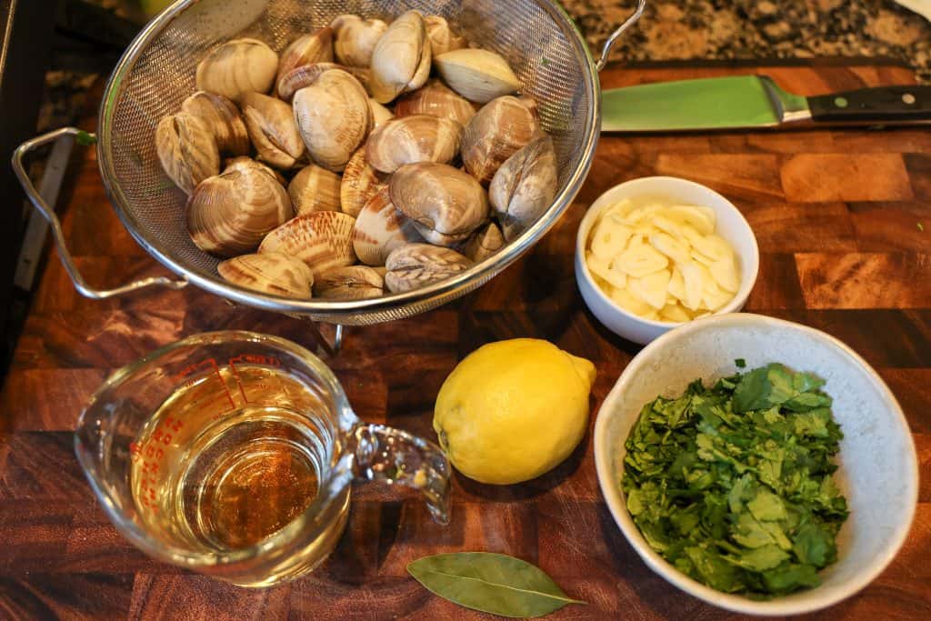 littleneck clams in a colander, vinho verde in a measuring cup, a whole lemon, bay leaf, sliced garlic, and fresh chopped cilantro on a wooden cutting board