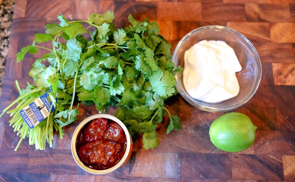 a bunch of cilantro, cup of greek yogurt, lime, and can of chipotles in adobo sauce on a wooden cutting board