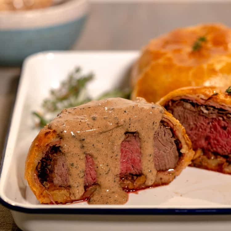Individual beef wellington cut in half with a black peppercorn sauce dripping down the center on a white baking sheet with thyme sprigs in background