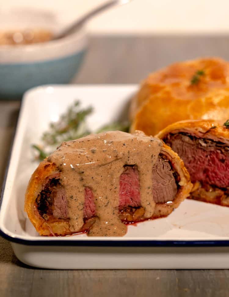 Individual beef wellington cut in half with a black peppercorn sauce dripping down the center on a white baking sheet with thyme sprigs in background