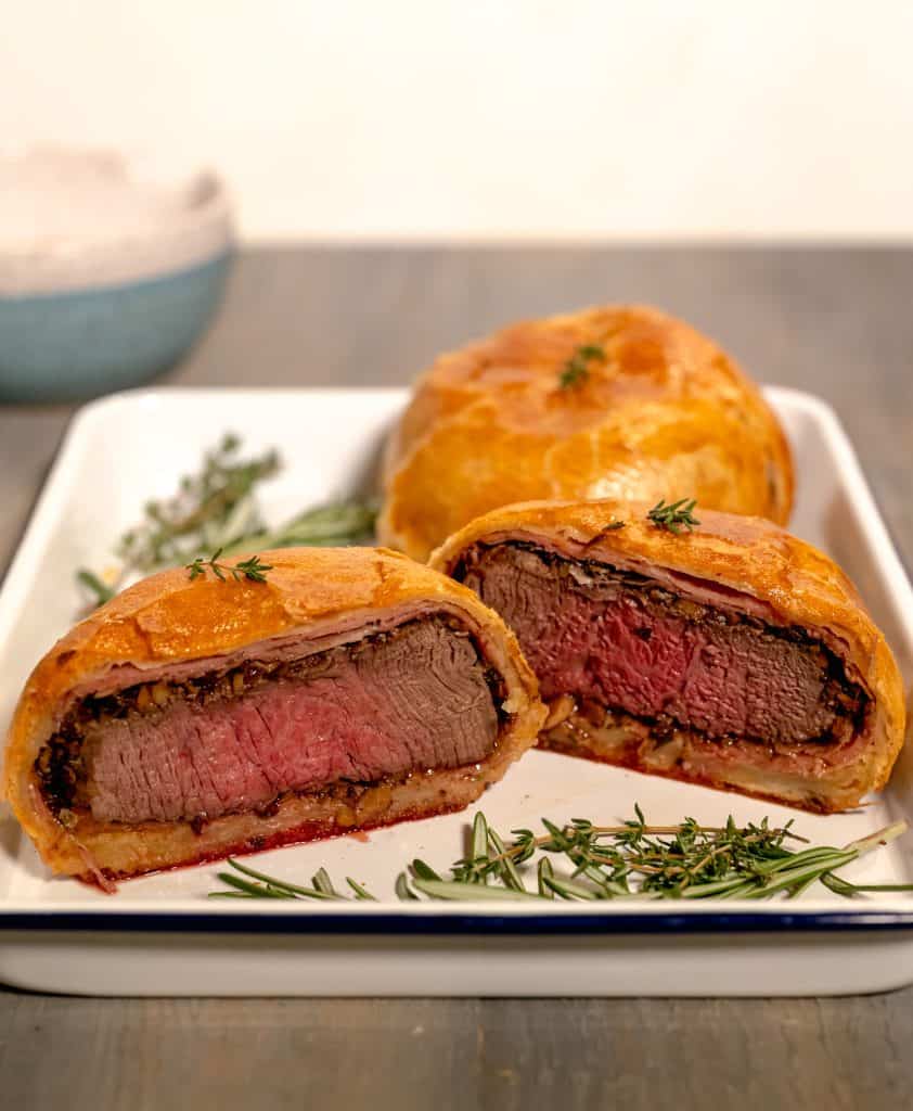 individual beef wellington sliced down the middle sitting on a white sheet pan with sprigs of thyme in the grey background