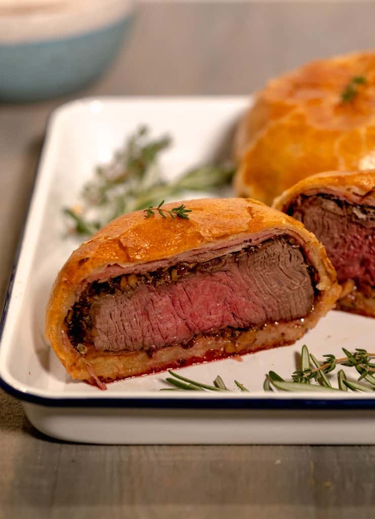 individual beef wellington sliced down the middle sitting on a white sheet pan with sprigs of thyme in the grey background 