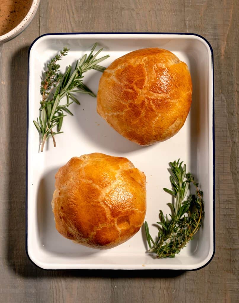 overhead shot of two individual beef wellingtons on a white sheet pan surrounded by fresh rosemary and thyme sprigs