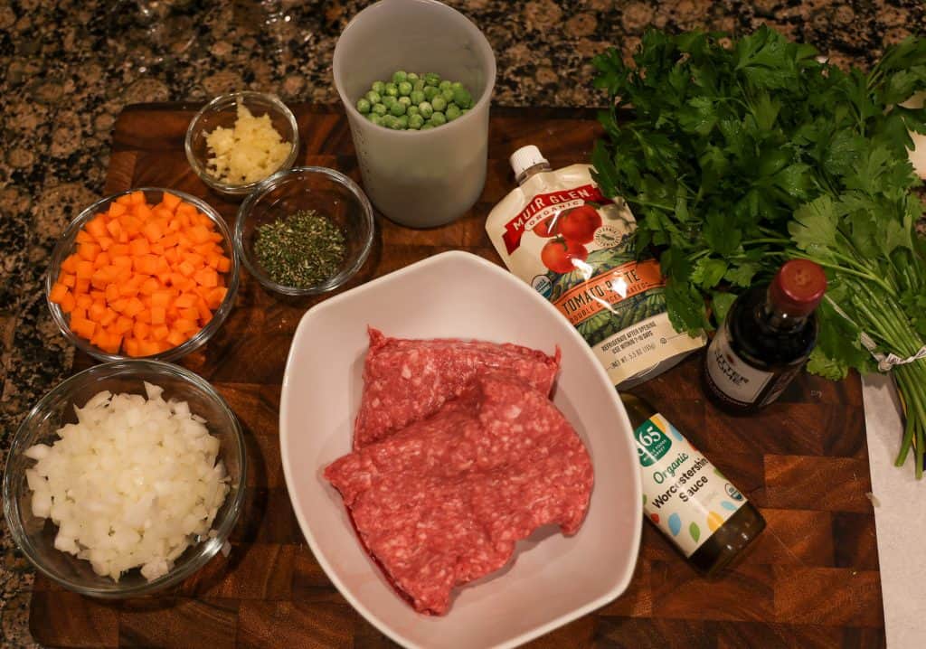 ground lamb in a white bowl, diced carrots and onion in a glass bowl, green peas, tomato paste, parsley, all displayed on a wooden cutting board.
