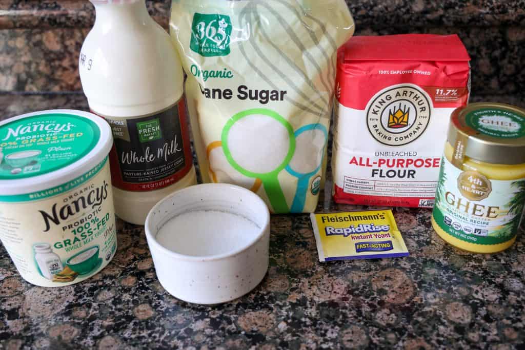 ingredients to make naan on the kitchen counter: bag of flour, bag of sugar, bottle of whole milk, carton of yogurt, ghee, packet of yeast, and kosher salt in a white bowl