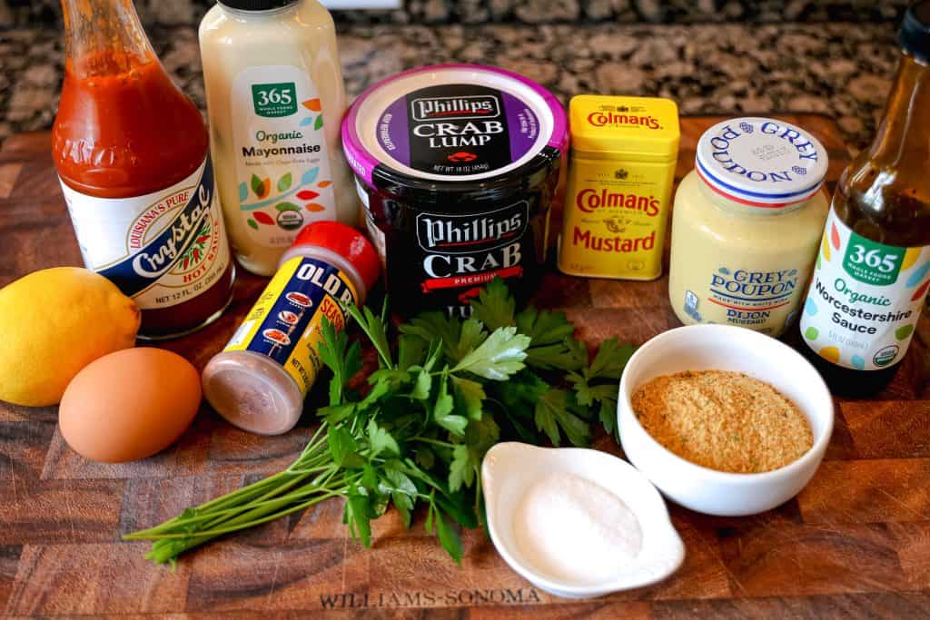 crab cake ingredients on a wooden cutting board: lump crab container, egg, breadcrumbs, fresh parsley, dijon mustard, hot sauce, lemon