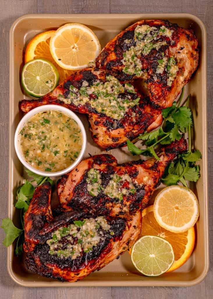 two grilled half chickens topped with cuban mojo sauce, surrounded by citrus slices, on a golden sheet pan