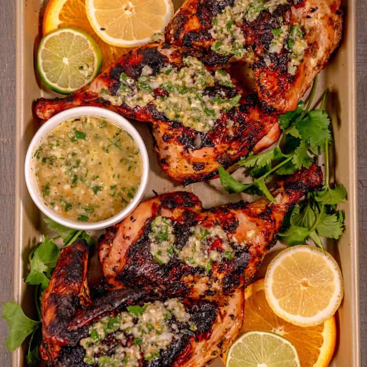 two grilled half chickens topped with cuban mojo sauce, surrounded by citrus slices, on a golden sheet pan