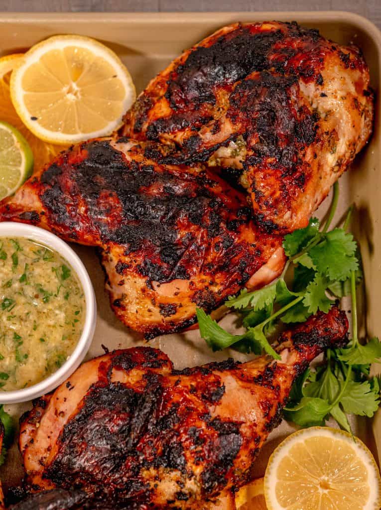 a grilled chicken halve on a gold sheet pan with slices of orange, lemon, and lime and a sprig of parsley surrounding