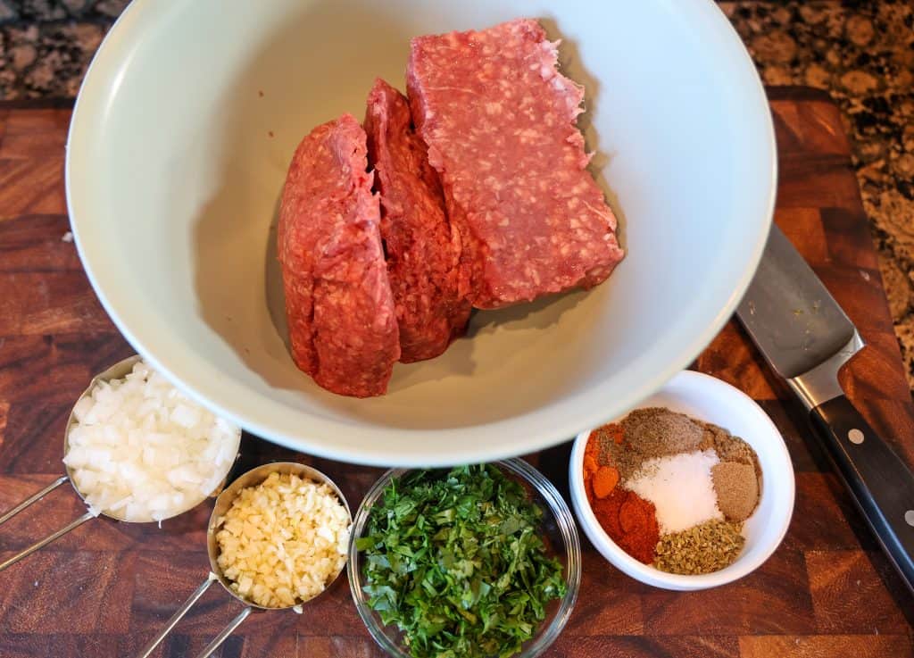 bowl filled with ground lamb and ground beef, chopped onion, garlic, parsley, and middle eastern spices on a wooden cutting board