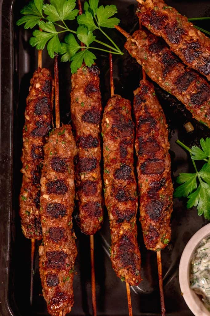 skewered beef and lamb koftas on a black sheet pan with a cup of tzatziki in a white bowl