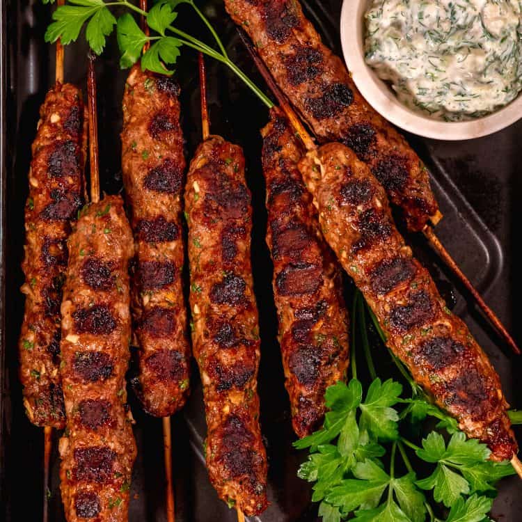 skewered beef and lamb koftas on a black sheet pan with a cup of tzatziki in a white bowl