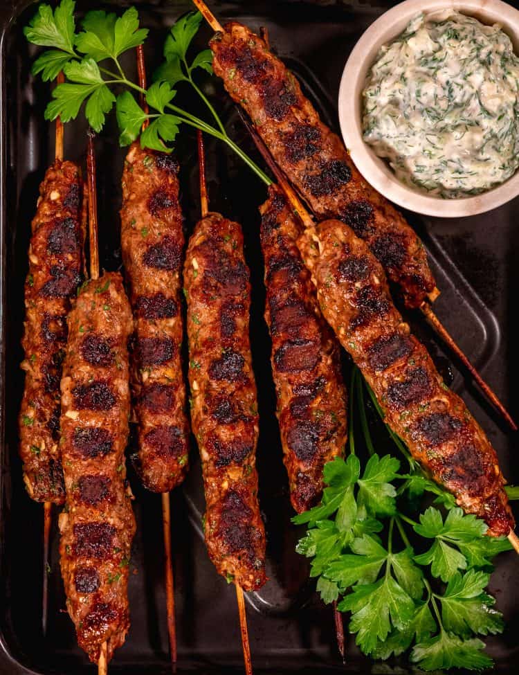 skewered beef and lamb koftas on a black sheet pan with a cup of tzatziki in a white bowl