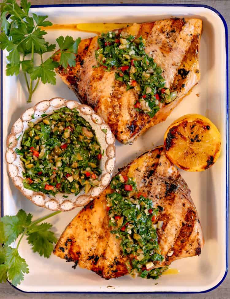a white sheet pan with two swordfish steaks topped with olive chimichurri. Two grilled lemon halves and a cup of chimichurri surrounds the swordfish steaks.