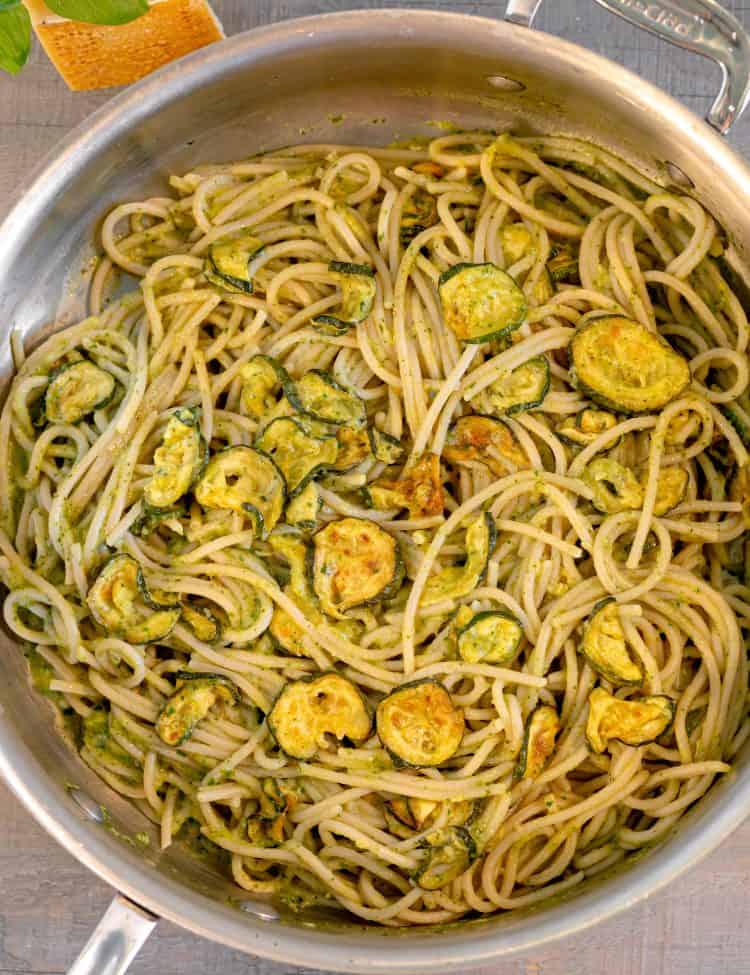 a skillet with pasta filled with zucchini coins on a grey background with a block of parmesan in the back