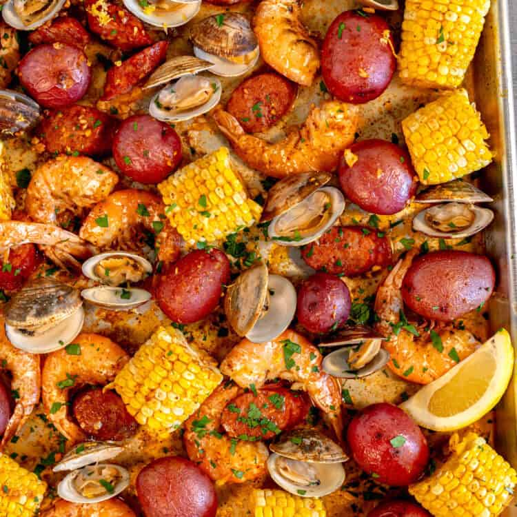 large sheet pan on a wooden surface filled with corn, red potatoes, sliced andouille sausage, clams, and pink roasted shrimp
