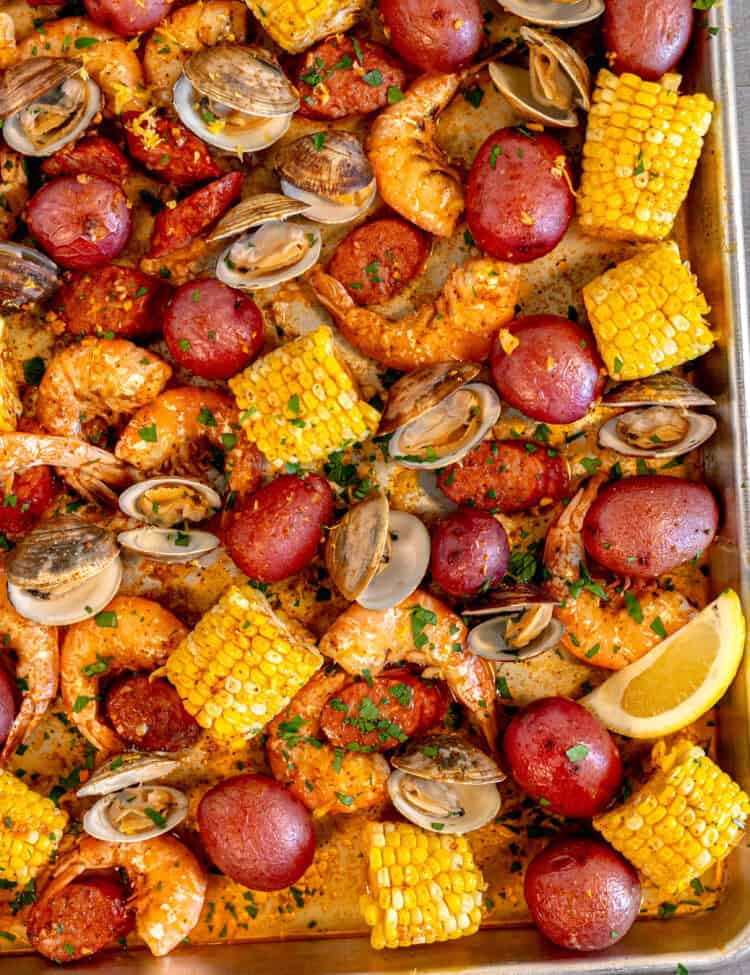large sheet pan on a wooden surface filled with corn, red potatoes, sliced andouille sausage, clams, and pink roasted shrimp