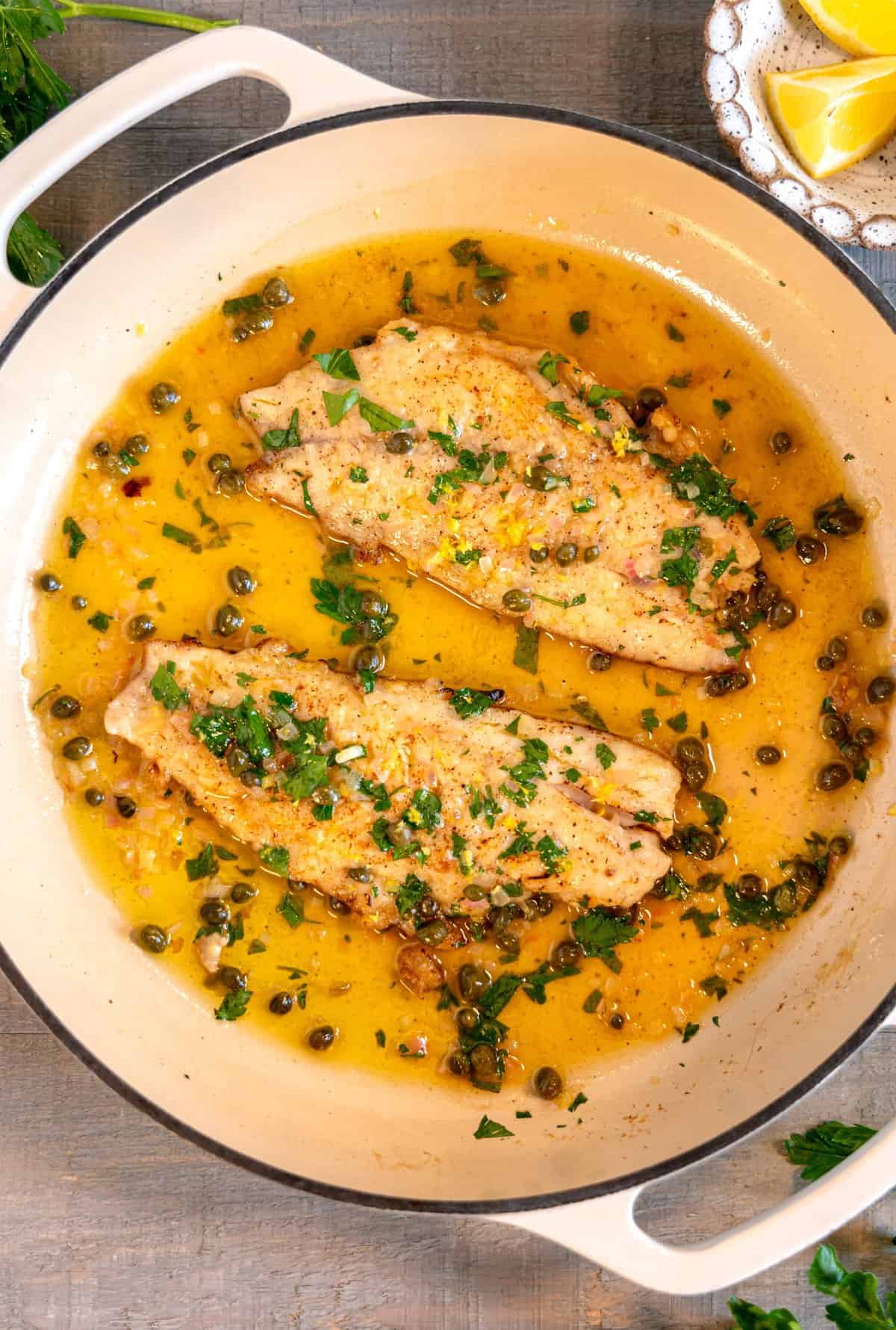 overhead two snapper fish filets in a white skillet covered in a buttery caper sauce