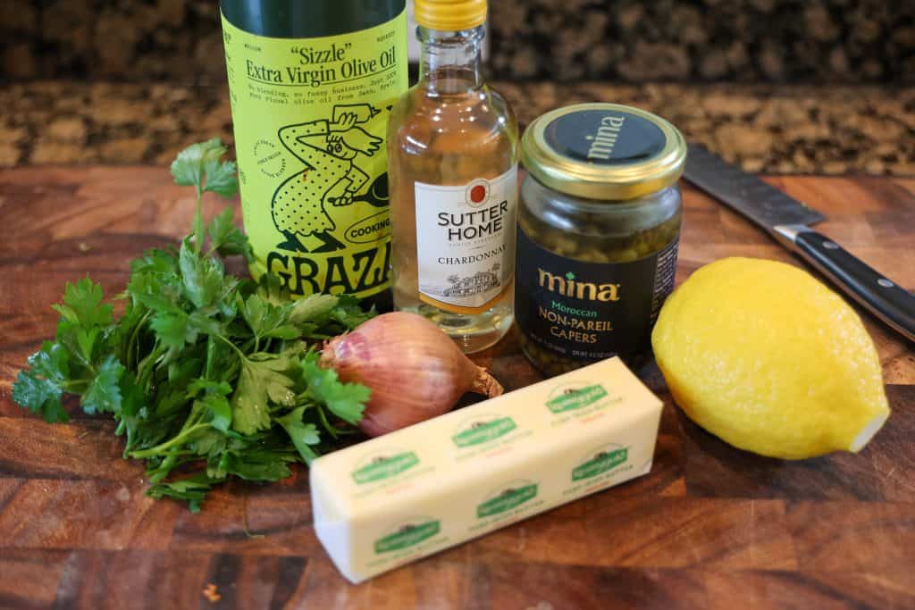 stick of butter, whole lemon, jar of capers, white wine, shallow, and parsley on a wooden cutting board