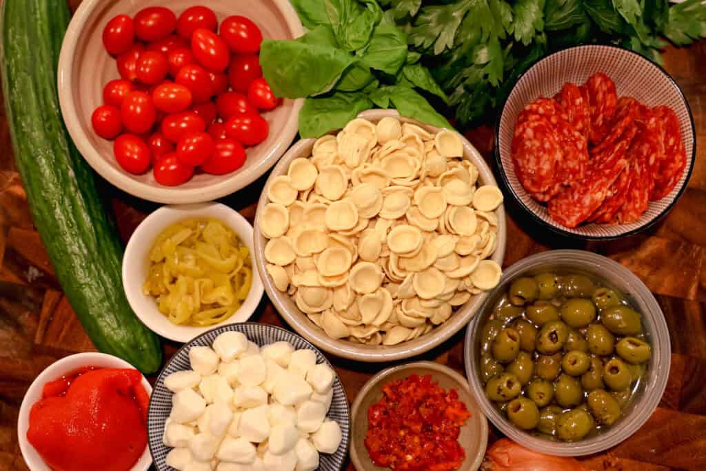 bowls of: dry pasta, olives, red peppers, tomatoes, mozzarella pearls, cucumber, and basil on a wooden cutting board