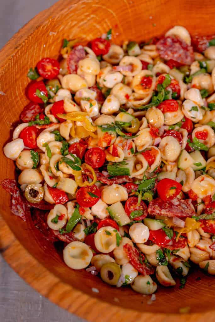 a wooden bowl on a grey surface filled with pasta salad of olives, pepperonis, tomatoes, basil, peppers, and mozzarella pearls