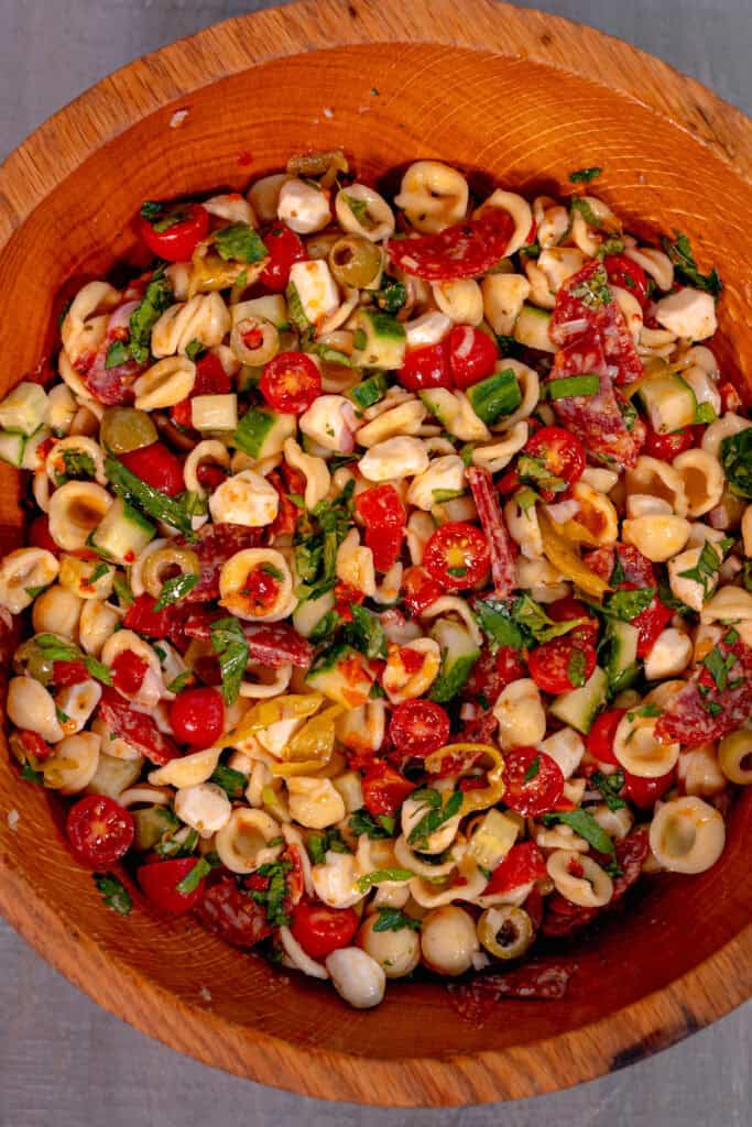 a wooden bowl on a grey surface filled with pasta salad of olives, pepperonis, tomatoes, basil, peppers, and mozzarella pearls