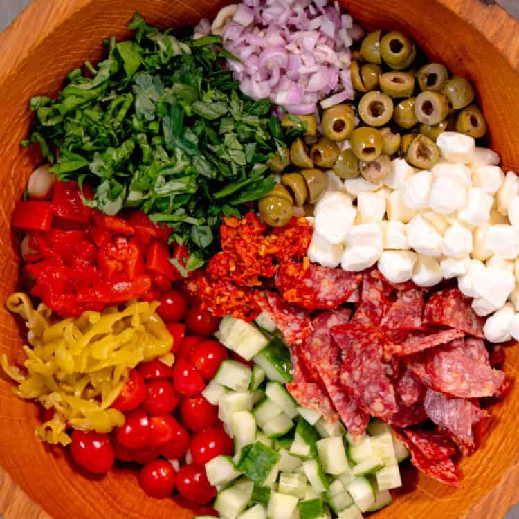 a wooden salad bowl filled with pepperonis, mozzarella, cucumbers, olives, red onion, parsley, basil, on a grey surface