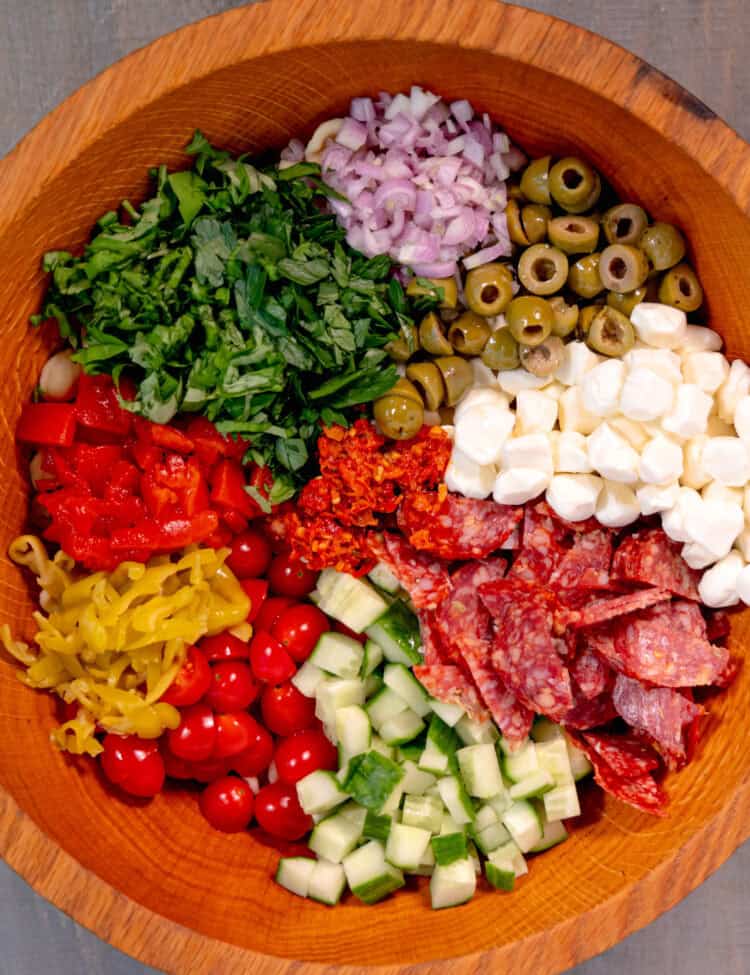 a wooden salad bowl filled with pepperonis, mozzarella, cucumbers, olives, red onion, parsley, basil, on a grey surface
