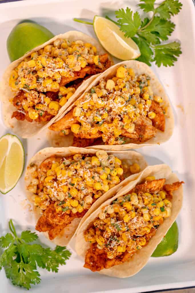 overhead photo of four chicken tacos topped with street corn elote topping surrounded by fresh cilantro and lime wedges