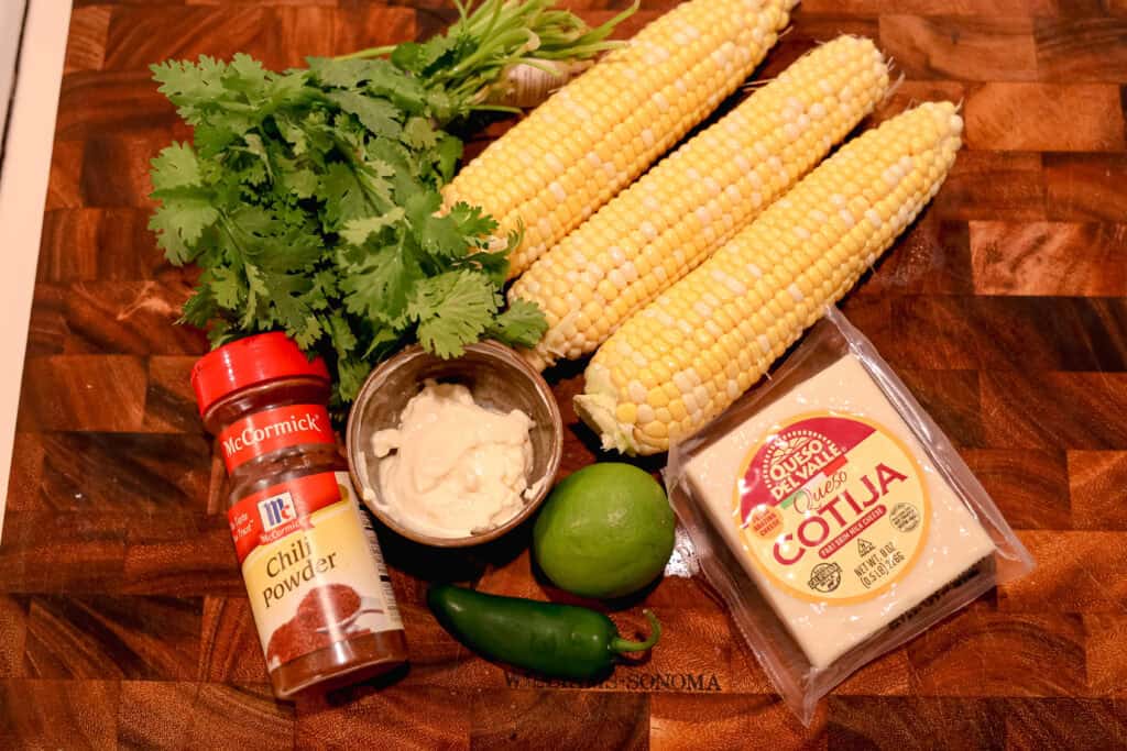 3 ears of corn, pack of cotija cheese, bushel of cilantro leaves, a lime, a jalapeno on a wooden cutting board