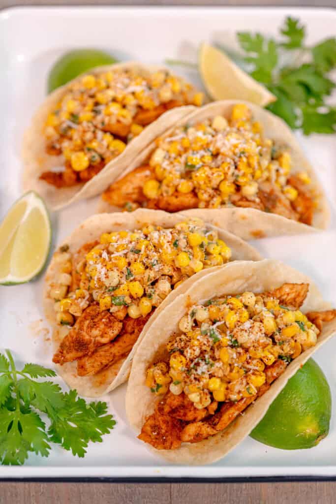 a white tray with four tacos filled with chicken and fresh corn surrounded by cilantro leaves and lime wedges