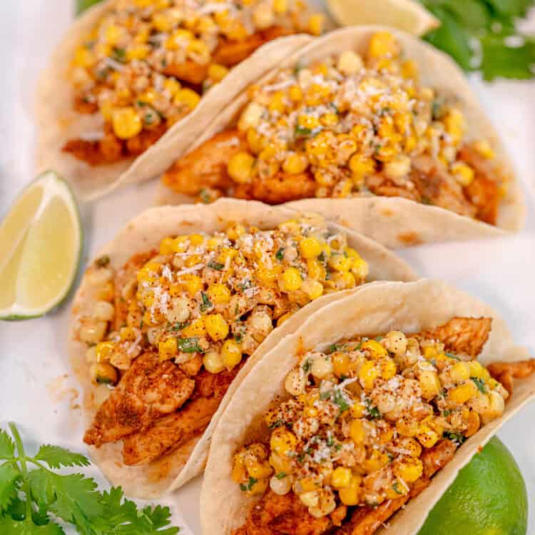 a white tray with four tacos filled with chicken and fresh corn surrounded by cilantro leaves and lime wedges