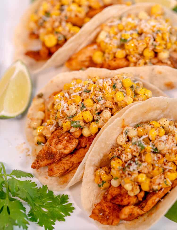 a white tray with four tacos filled with chicken and fresh corn surrounded by cilantro leaves and lime wedges