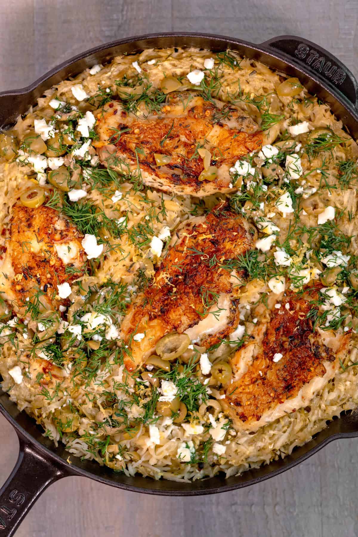 cast iron skillet on a gray wooden background, filled with rice topped with chicken breast, feta cheese, and fresh dill