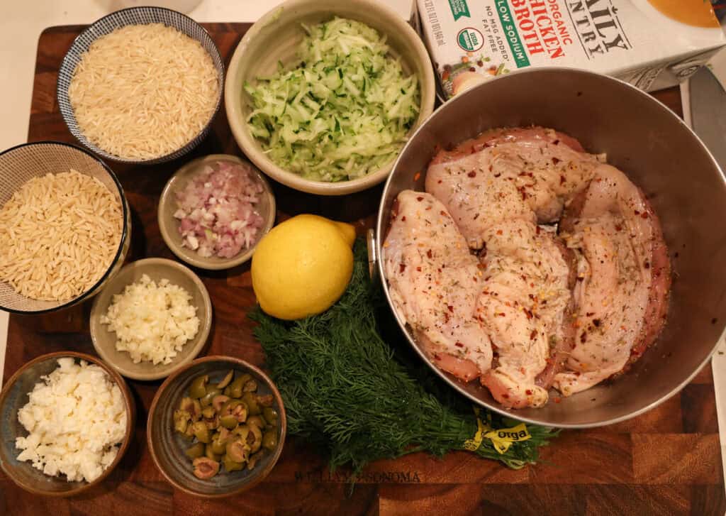 bowl of chicken, shredded zucchini, basmati rice, orzo, minced shallot, minced garlic, chopped olives, bunch of dill, and lemon on a wooden cutting board